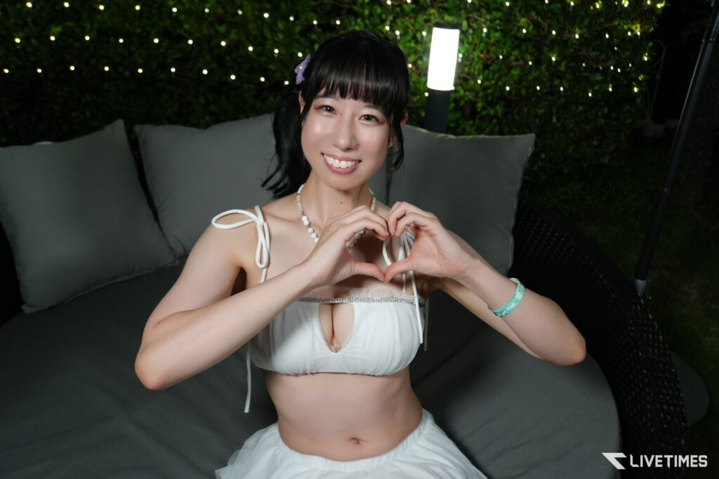 Smiling woman in a white outfit on an outdoor couch at night, forming a heart with her hands, fairy lights in the background.