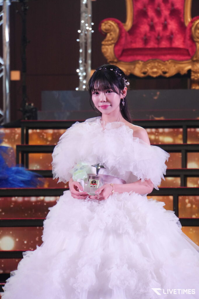 Young woman in a white, voluminous ruffled gown on stage, holding a glass trophy in front of an ornate gold throne backdrop.