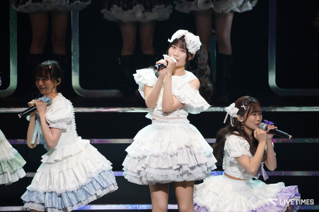 Female pop idols in white frilly outfits performing on stage with microphones, mid-song pose.