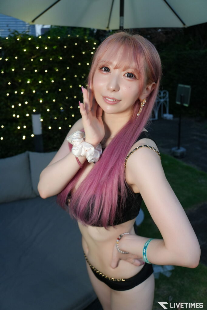 Woman with pink hair posing for a selfie in a black bikini top outdoors under a large umbrella, string lights in the background.