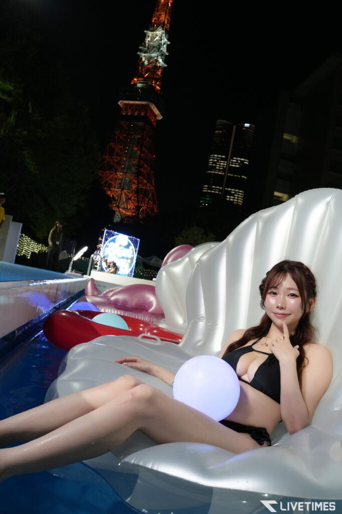Young woman in a black swimsuit sits in a white inflatable raft on a water ride at night, with a tall illuminated tower in the background.