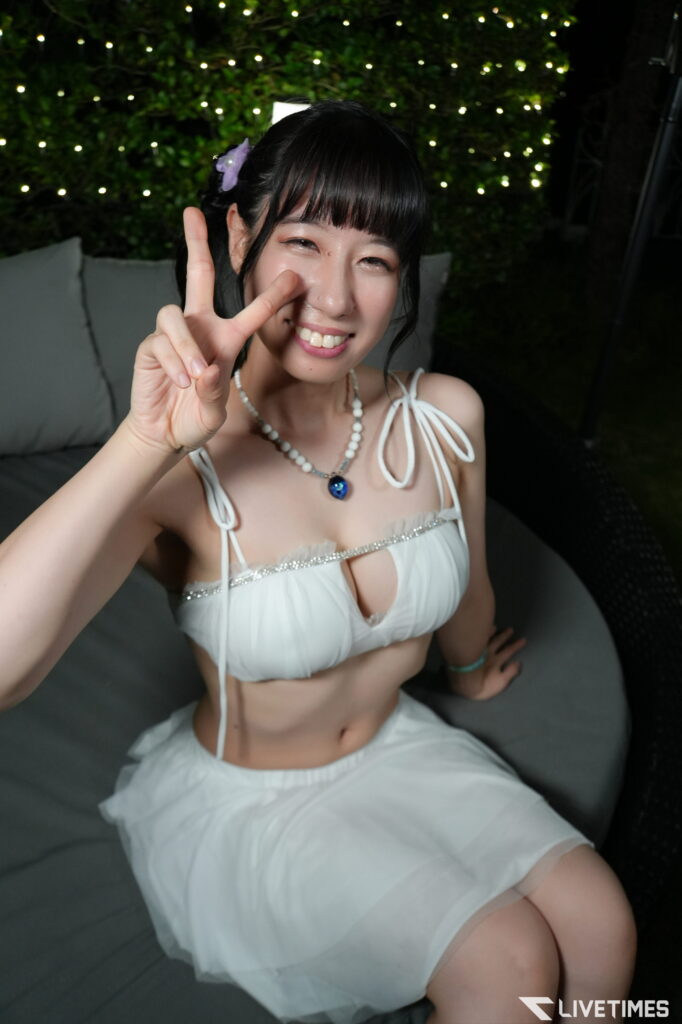 Smiling young woman in a white outfit makes a peace sign at the camera, with string lights in the background.