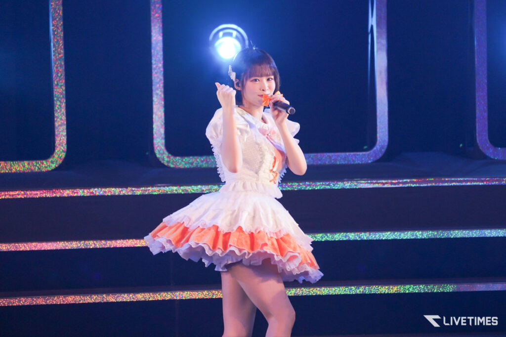 Female singer in a white pink frilly dress performs on stage with a microphone and a glittery backdrop behind her.