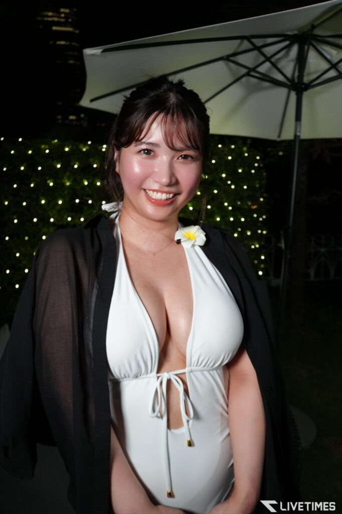 Smiling woman in a white halter swimsuit with a black sheer cover-up, standing at night with a large patio umbrella and twinkling background lights.
