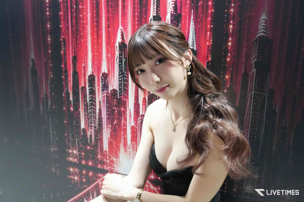 Portrait of a woman with long wavy hair in a black strapless top, posing against a red neon cityscape backdrop (LIFETIME S watermark visible).