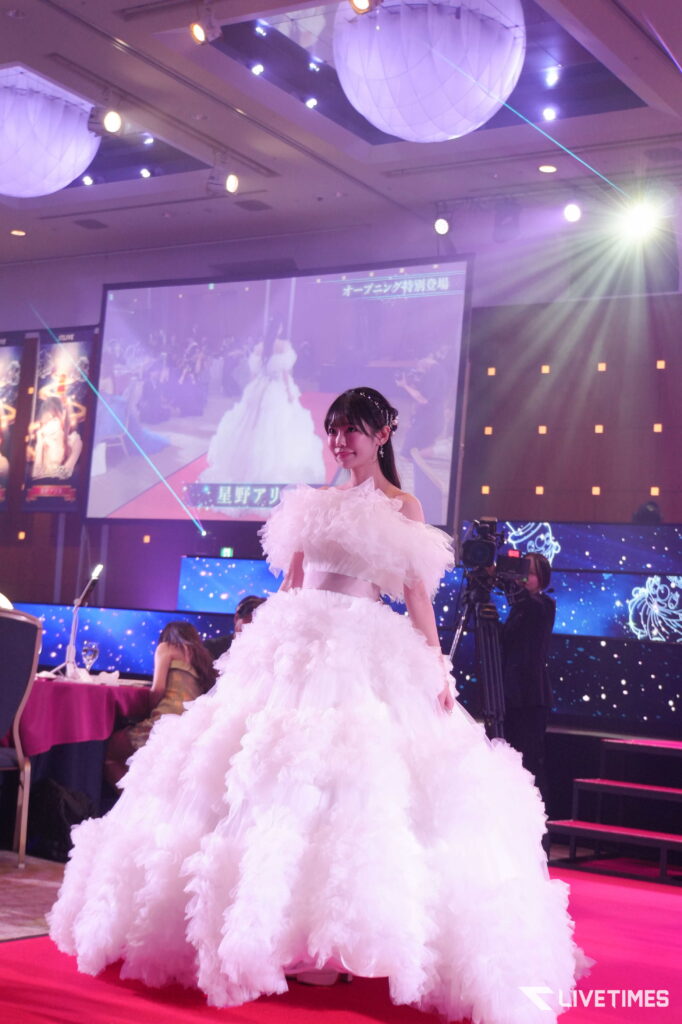 Young woman in a voluminous white gown posing on a pink-carpeted stage at a fashion/event with a large screen and colorful stage lights in the background.