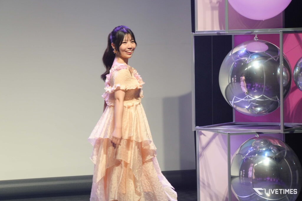 Young woman in a peach lace gown posing on stage beside a display of large reflective glass spheres.