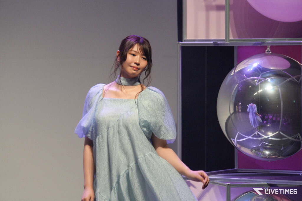 Young woman in a pale blue lace dress standing beside a large glass sphere exhibit with purple lighting in a showroom or event space.