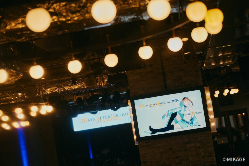 Indoor stage with warm round pendant lights, a brick pillar, and a large screen displaying a fashion photo and '2DAYS AFTER BIRTHDAY 2026'