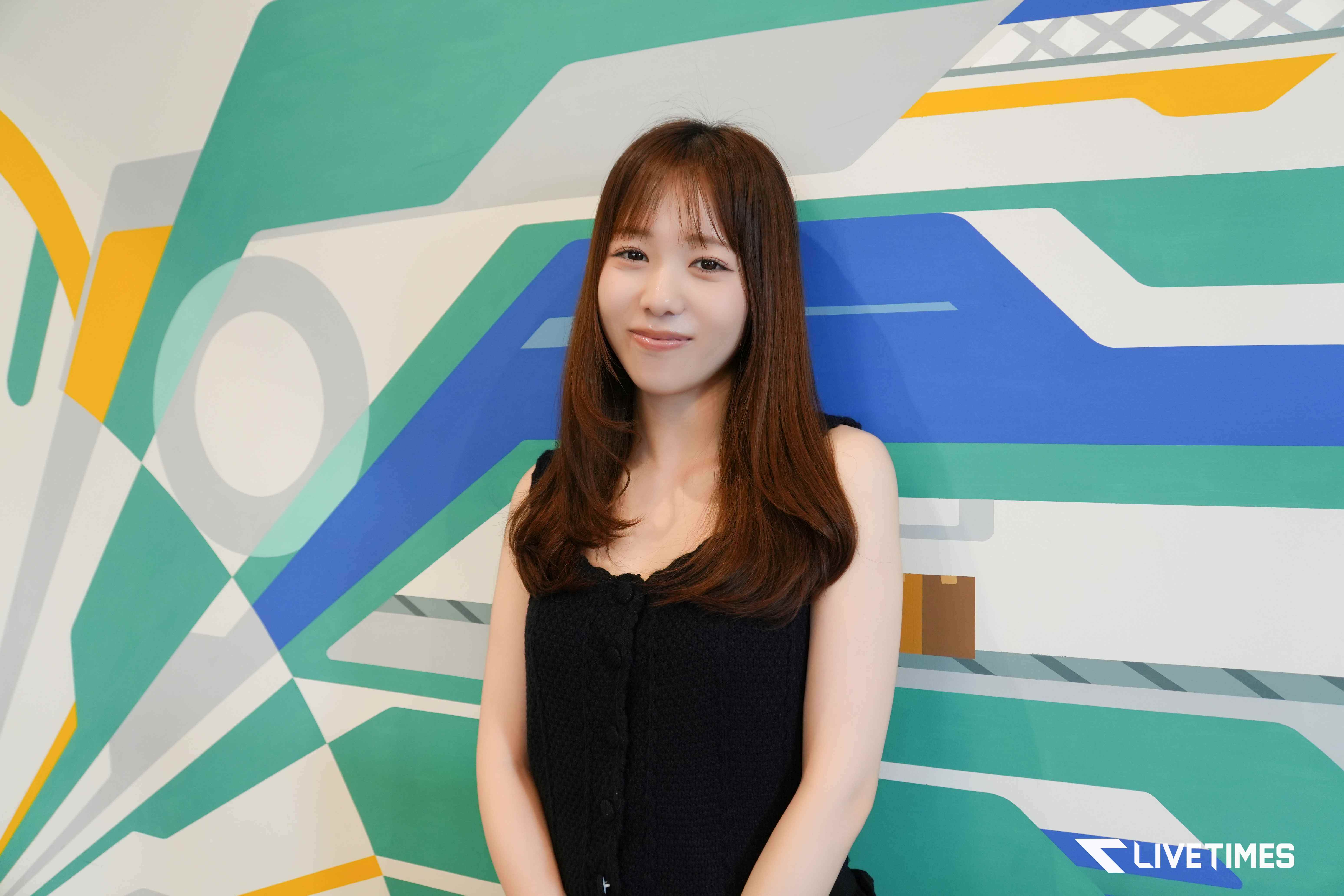 Portrait of a young woman with long brown hair in a black sleeveless top, standing before a colorful geometric mural.