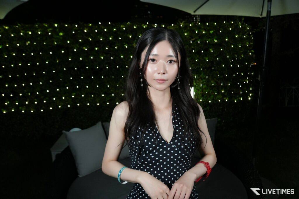 Young woman in a black polka-dot dress posing outdoors at night with string lights behind her.
