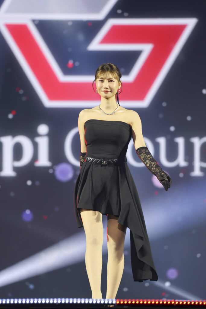 Female performer in a strapless black dress with lace gloves, standing on a lit stage with a large red-and-white geometric logo behind her, jewelry visible.