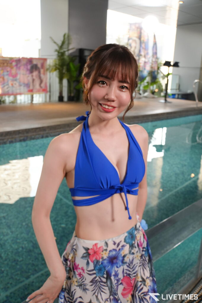 Smiling woman in a blue halter bikini top and floral skirt standing by an indoor pool.