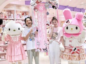 Two people in pink Hello Kitty outfits posing with oversized pink plush mascots in a pastel store setting.