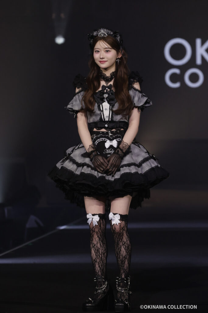 Young woman on a dark stage wearing a gray-and-black Gothic Lolita dress with lace gloves, bows, and a tiara, posing for the audience.