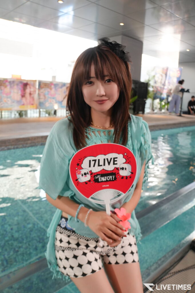 Young woman by an indoor pool in a turquoise top, holding a red 17LIVE fan with panda graphics.