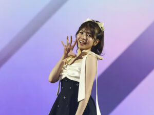 Smiling female performer with a cream bow in her hair, waving at the audience on stage with a purple backdrop.