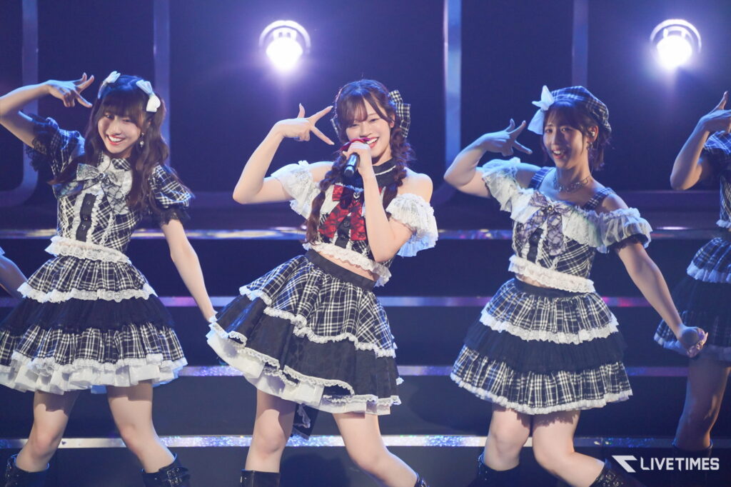 Group of female performers in matching plaid stage outfits singing and dancing under blue lights on stage.