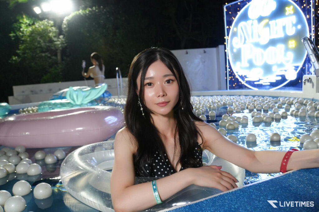Woman in a polka-dot swimsuit sits in a silver inflatable pool float at night, with a neon 'Night Pool' sign in the background.