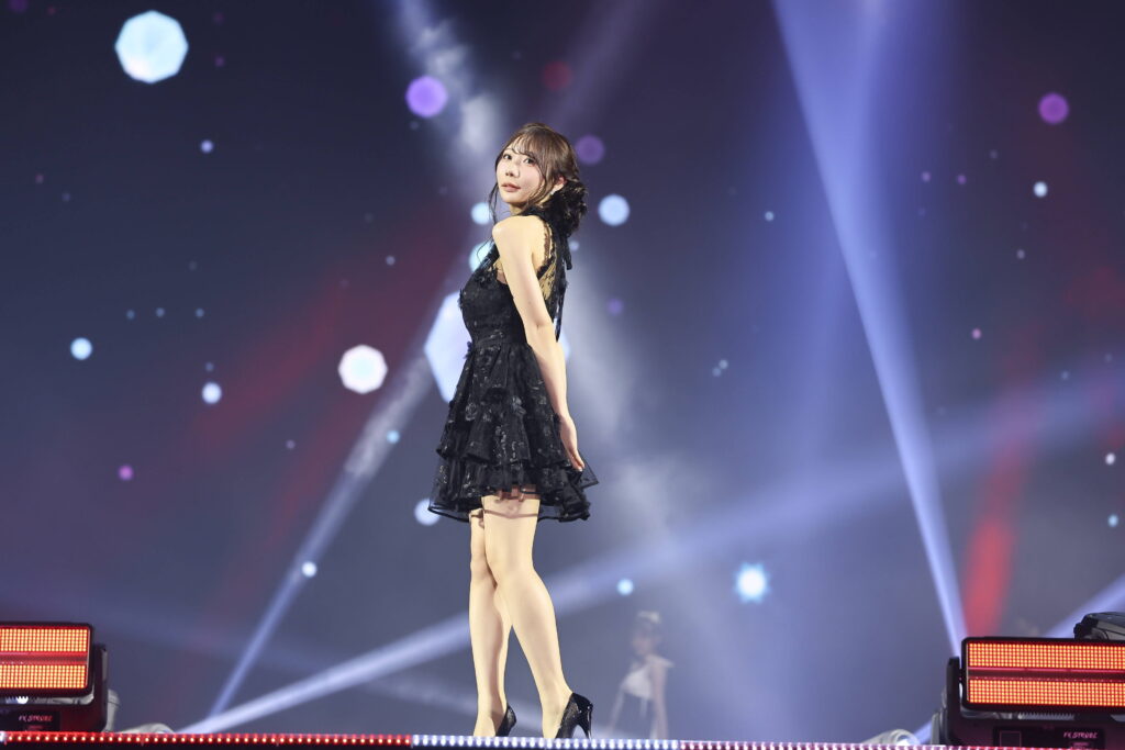 Female performer in a black lace dress standing on a lit stage, facing slightly to the left with colorful bokeh lights in the background.