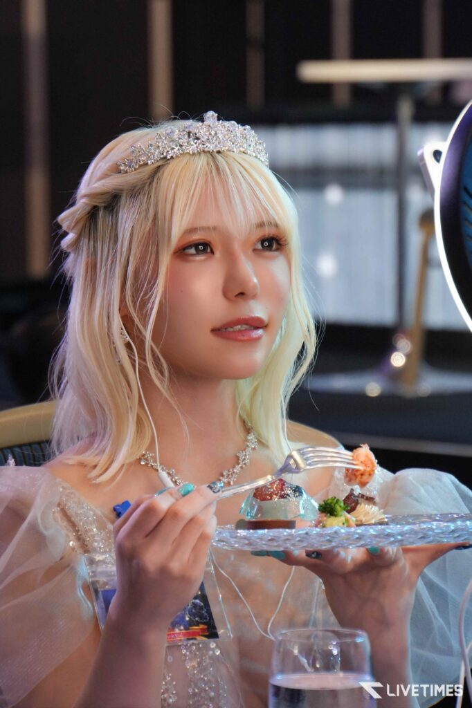Blonde woman wearing a jeweled tiara and pearl earrings enjoys a plated dessert with a fork at a formal event.