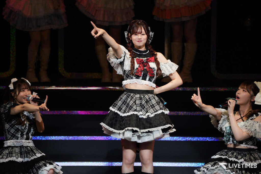 Center-stage idol in a black-and-white plaid costume with lace, pointing upward during a live performance while surrounded by backup dancers holding microphones.