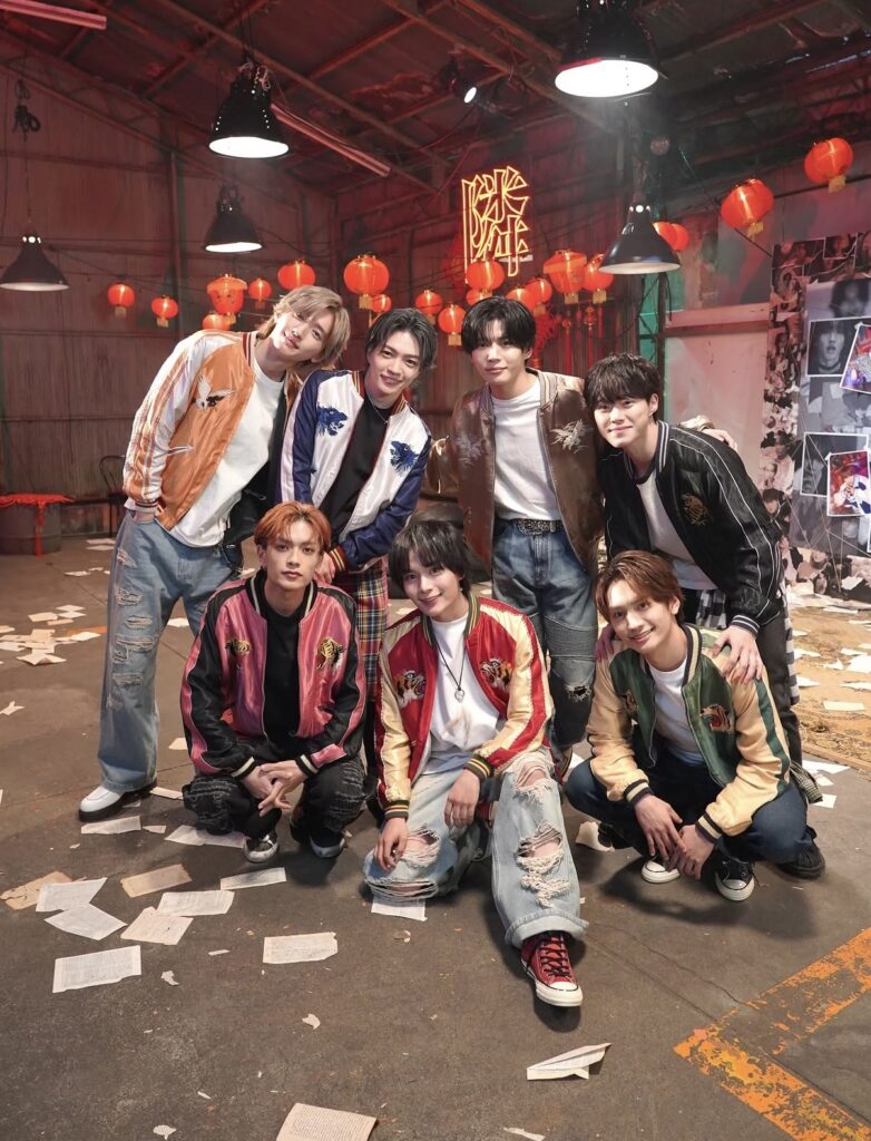 Seven young men in colorful bomber jackets posing in a studio with red lanterns and wall collages nearby.
