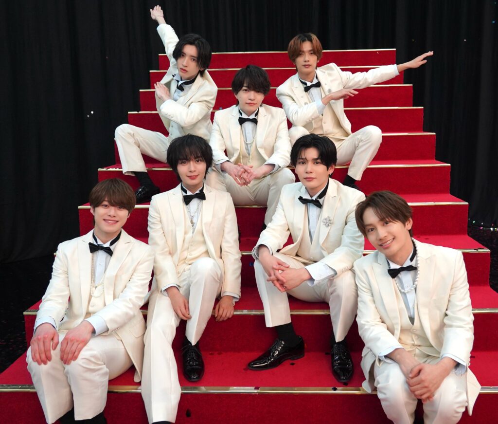 Group of young men in matching white tuxedos posing on a red-carpet staircase, some seated and others standing, smiling for a team photo in front of a black backdrop.