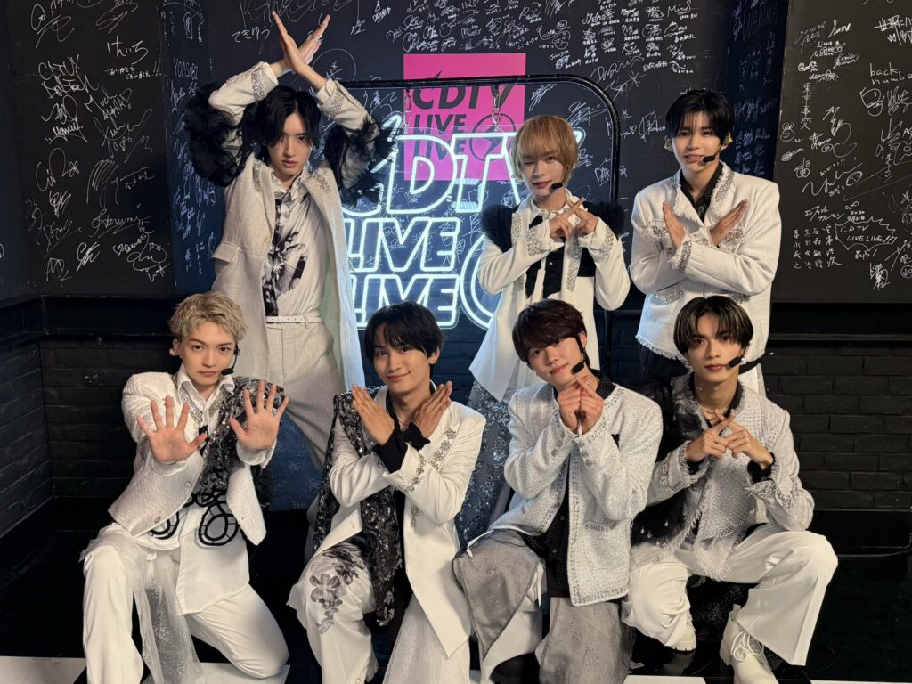 Eight members of a K-pop group pose together in white, sparkly outfits in a signed-wall backstage area with a neon backdrop showing 'LIVE' signs.
