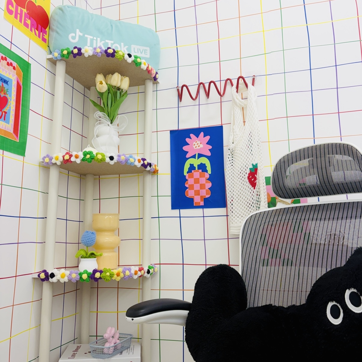 Playful office nook with a pastel TikTok Live pillow on a flower-decorated shelf and a vase of white tulips. - Colorful grid wall with a decorated shelf, flowers, and a blue poster featuring a pink flower and pineapple. - White mesh-backed office chair beside a red hook rack and a hanging tote bag on the wall. - Large black plush bear resting near a striped mesh chair in a bright, whimsical workspace. - Small pink bunny figurine in a basket on a lower shelf under the flower garland.