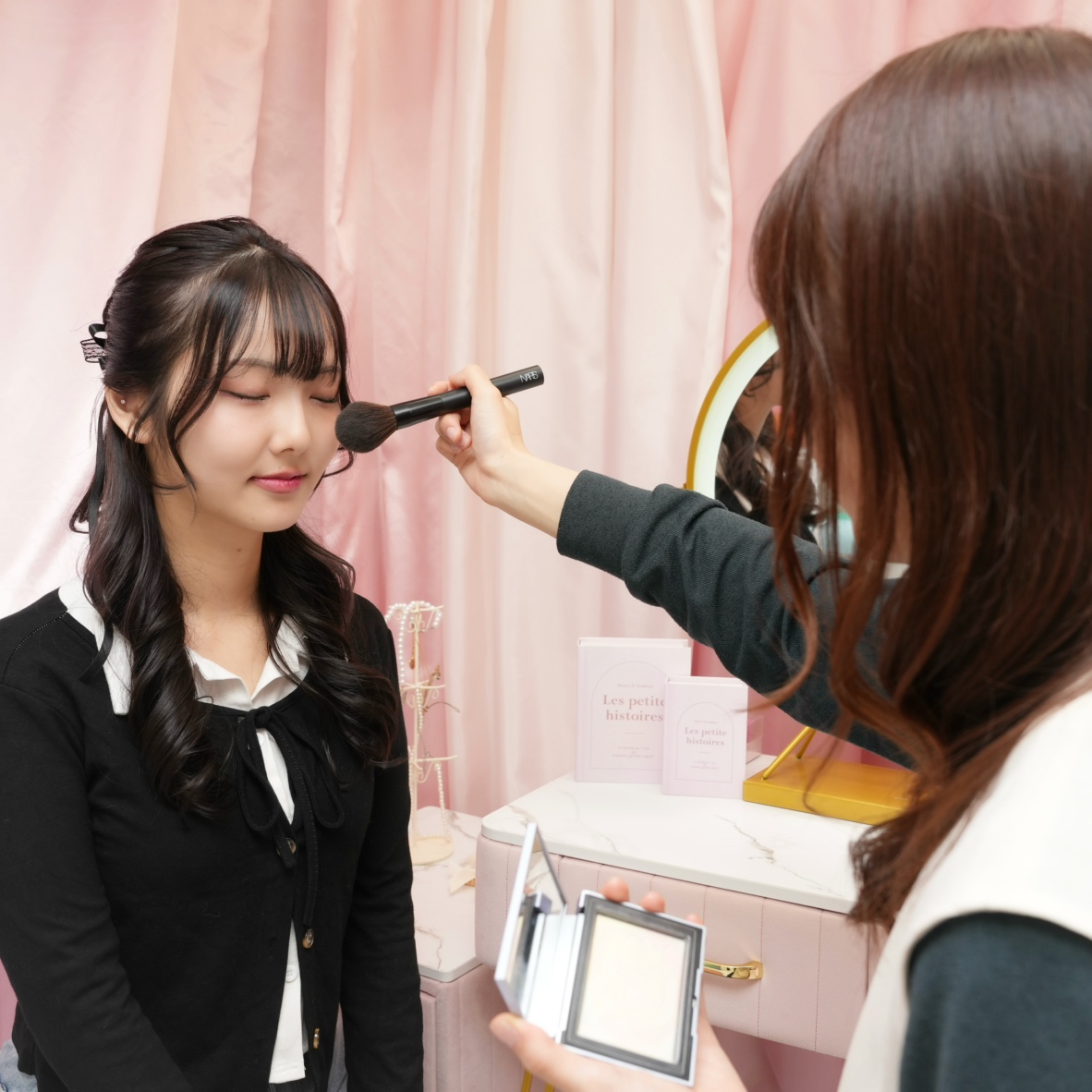 Young woman with eyes closed as a makeup artist blends powder with a brush in a pink boutique setting.