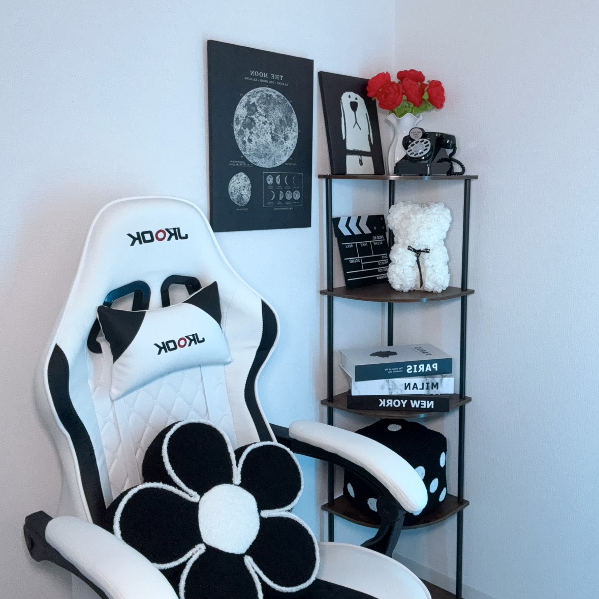 White and black gaming chair with flower-shaped pillow in a corner study setup, next to a shelving unit with decor and books.