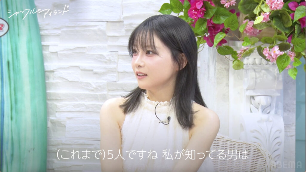 Young woman with black hair in a light beige top gazes to the left, pink flowers and a white brick wall behind her.