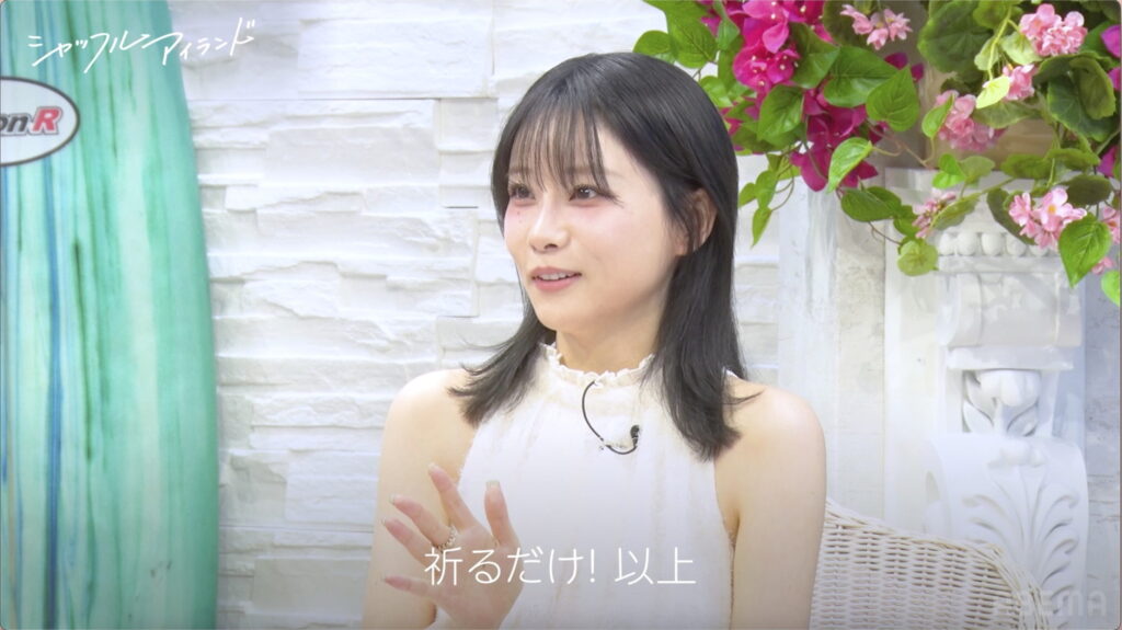 Woman with dark hair in a white sleeveless top sits in a pastel studio setting with pink flowers and a teal column to the left. Japanese text overlays the scene.