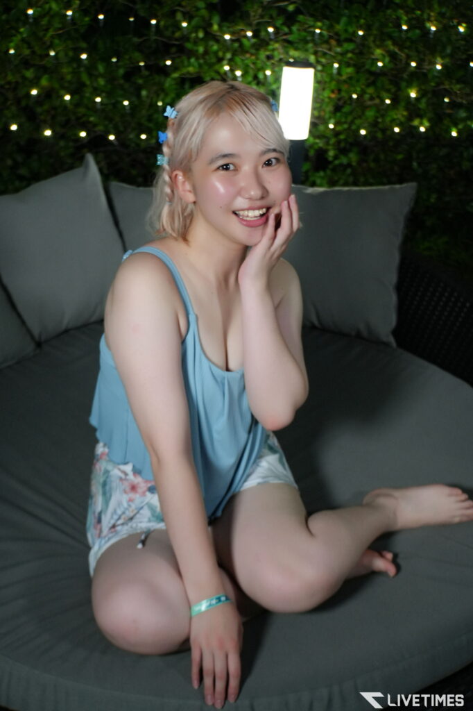 Young woman with short blond hair in a light blue tank top sits on an outdoor sofa, smiling with a hand to her cheek; string lights glow in the background.