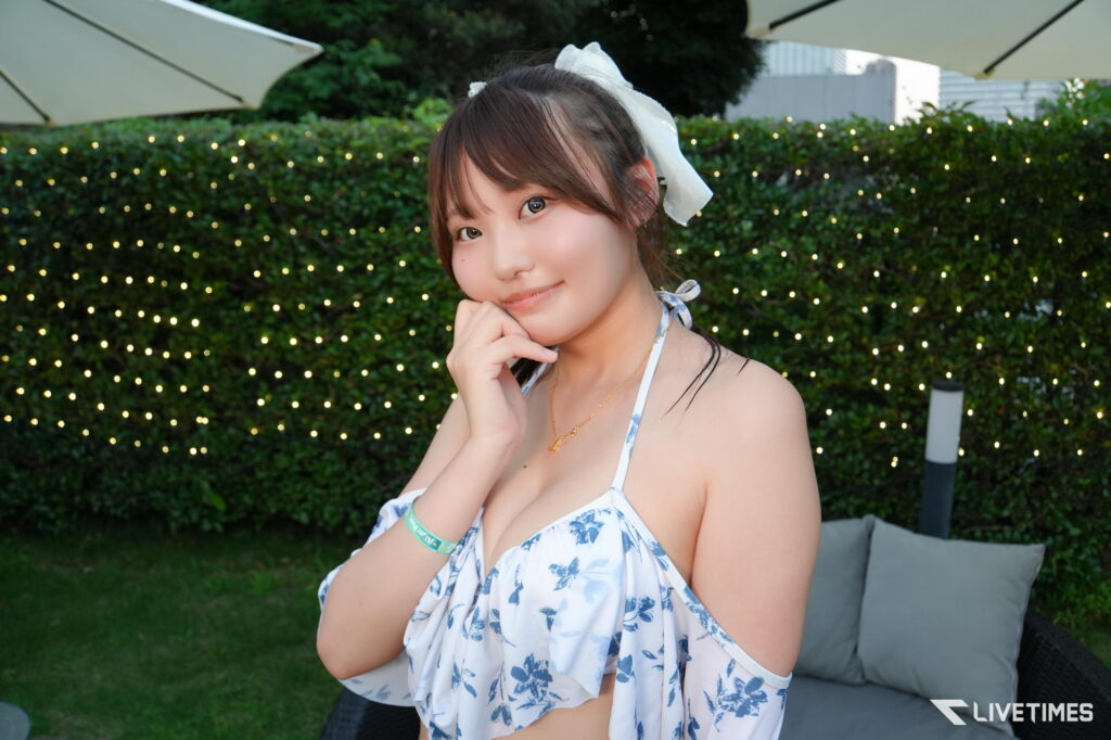Woman posing outdoors in a white and blue floral top with a white hair bow, hand under chin, string lights in the background.