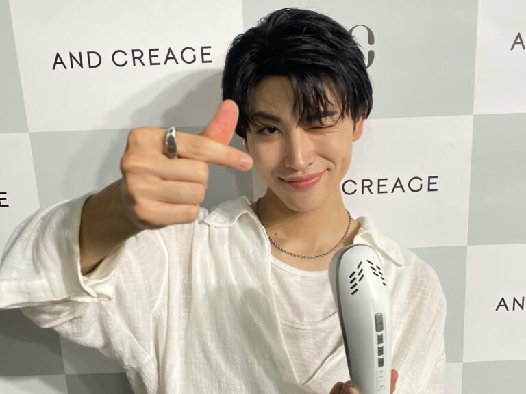 Person in a white shirt smiling at the camera while making a finger-heart gesture with their left hand, holding a white microphone against a tiled backdrop.