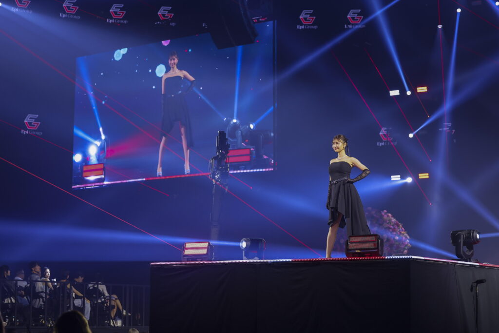 Model in a black gown posing on a brightly lit stage with a large screen showing her reflection in the background, an audience visible in front.