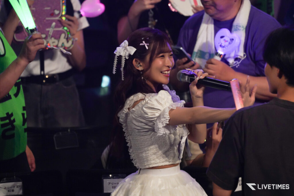 Young woman in a white frilly dress singing into a microphone at a fan event, smiling at the crowd surrounded by supporters with glow sticks