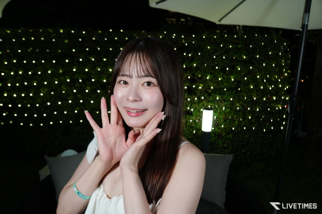 Young woman smiling at the camera, hands raised near her face, nighttime backdrop of twinkling lights in the background.