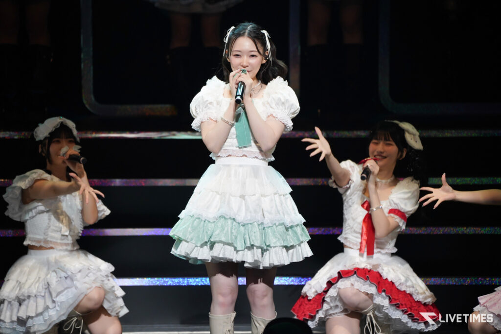 Center-stage singer in a mint-green layered dress sings into a microphone, flanked by two dancers in white frilly outfits on a concert stage.