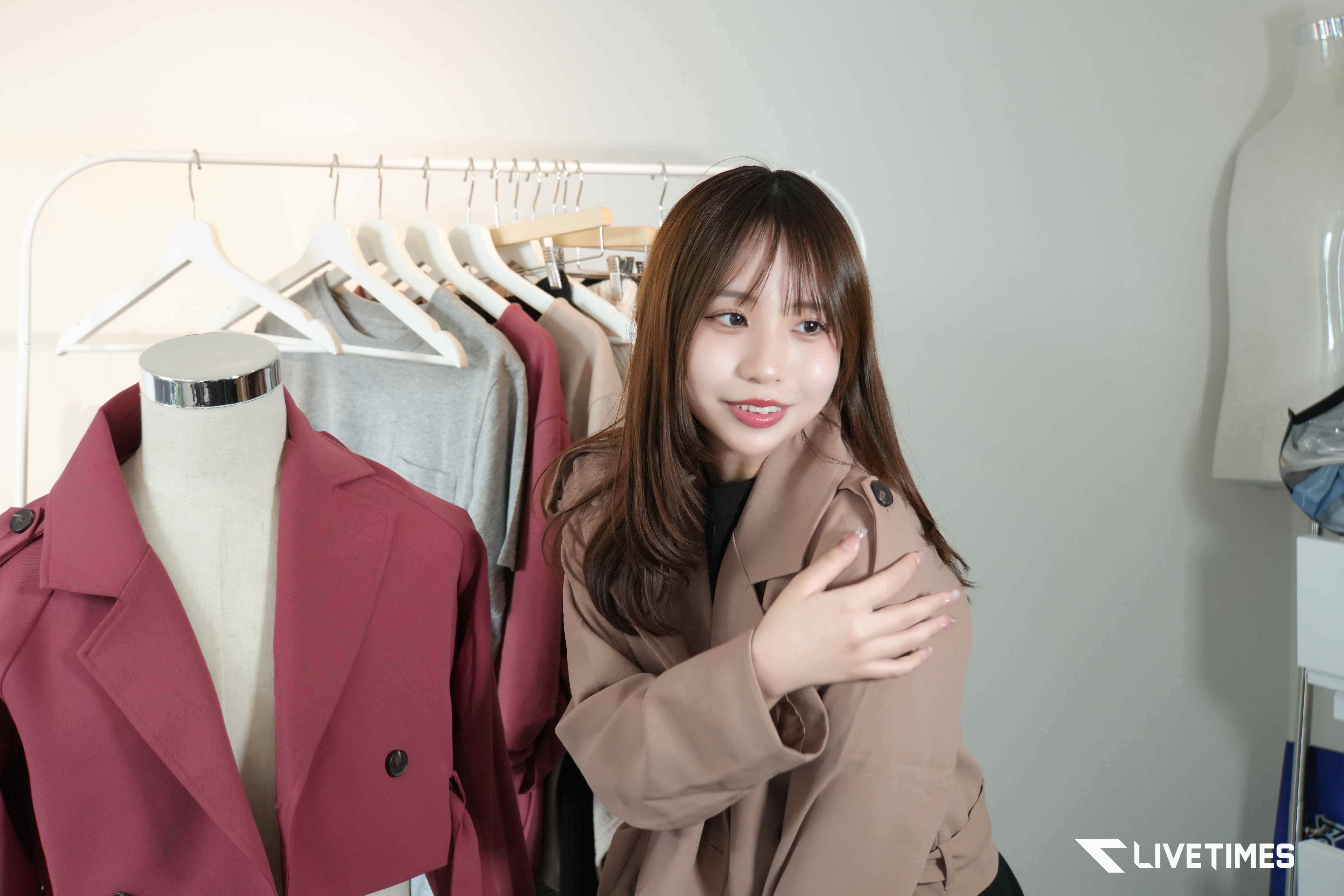 Woman trying on a beige trench coat in a clothing store, with a rack of coats and a mannequin beside her.