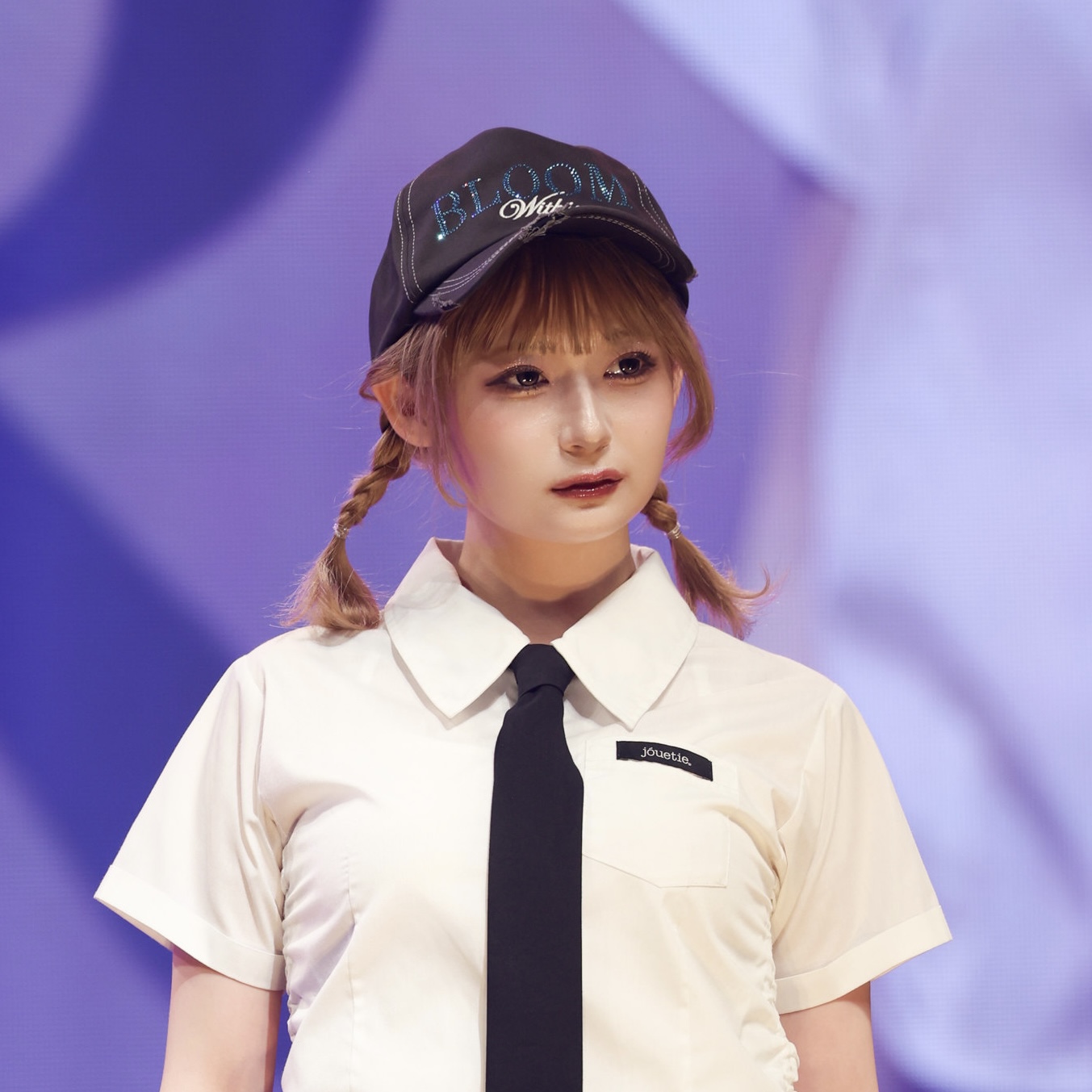 Young woman with braided pigtails wearing a white short-sleeve shirt, black tie, and a cap that reads BLOOM on a purple stage backdrop.