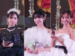 Three women in formal gowns hold trophies on stage, each smiling at the awards ceremony (left in black lace, center in white ruffles, right in white off-shoulder).