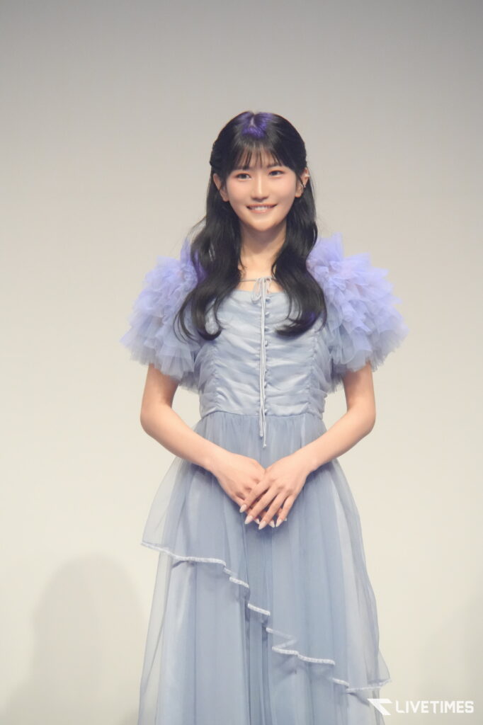 Smiling young woman in a light blue, ruffled-sleeve dress standing against a plain light background, hands gently clasped in front.