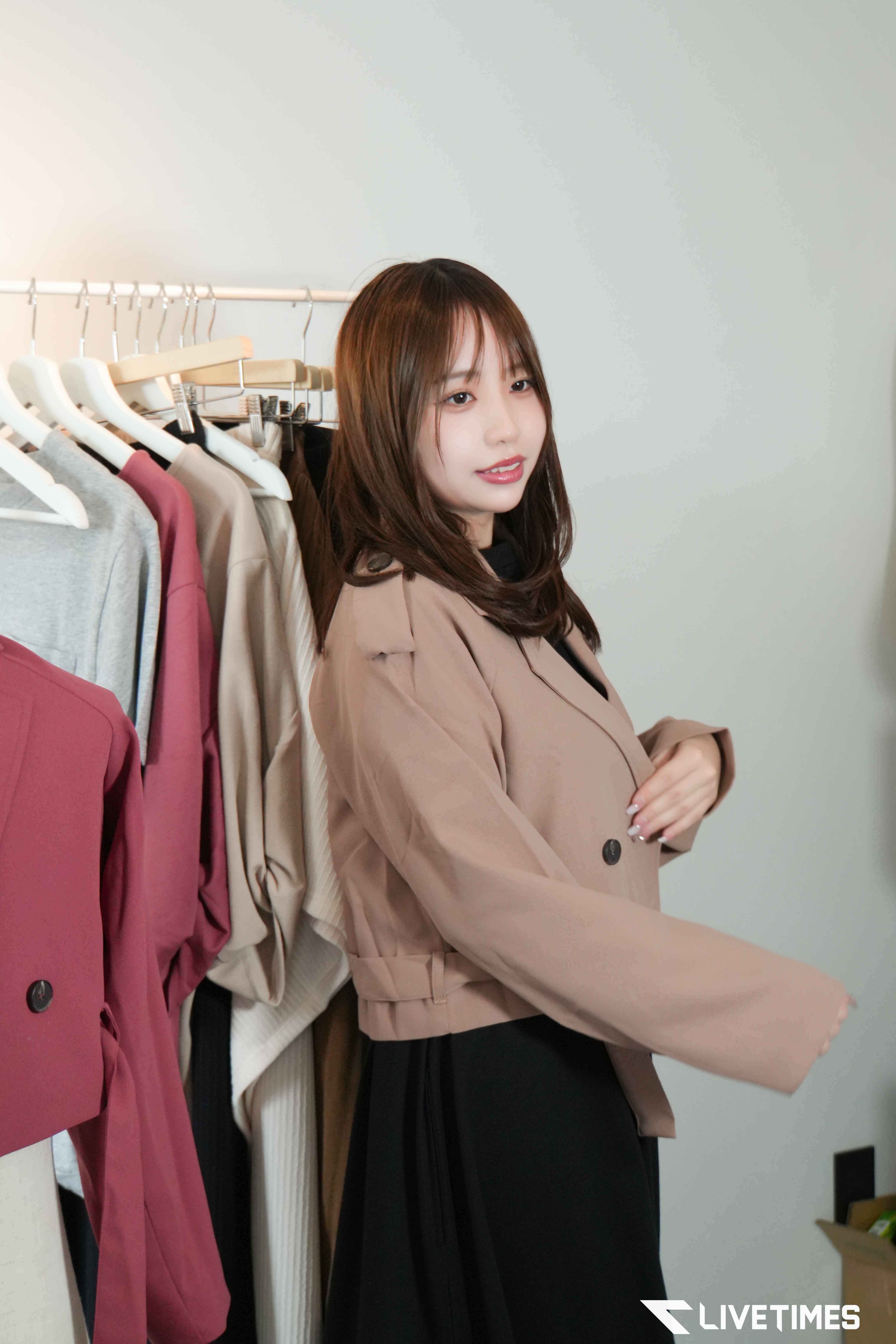 Woman in a beige trench coat standing beside a clothing rack with colorful jackets on hangers in a boutique setting.