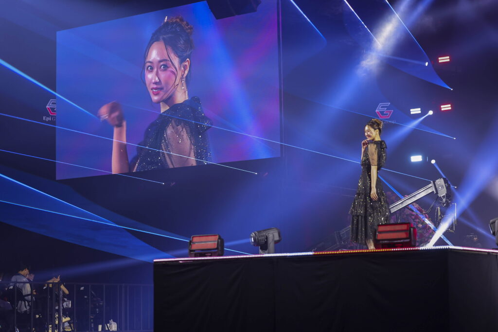 Singer on a glittery black dress performs on a stage with a large screen showing her close-up and blue laser lights surrounding her spotlit area, creating a concert atmosphere.