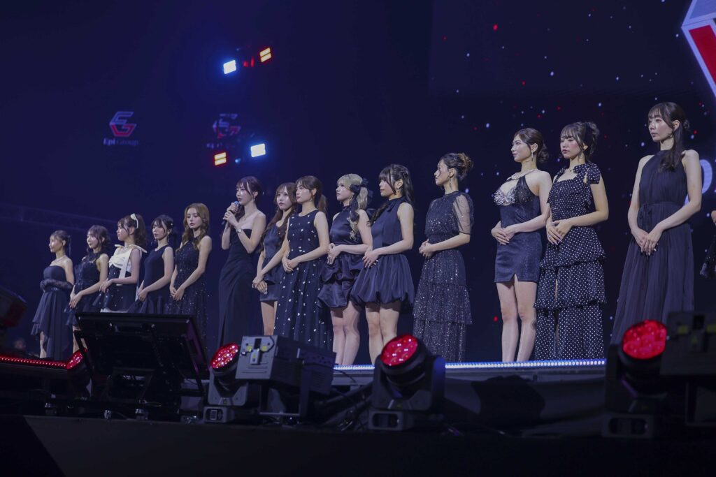 Group of female performers in navy and black evening gowns standing in a line on stage during a performance, blue stage lights in the background.