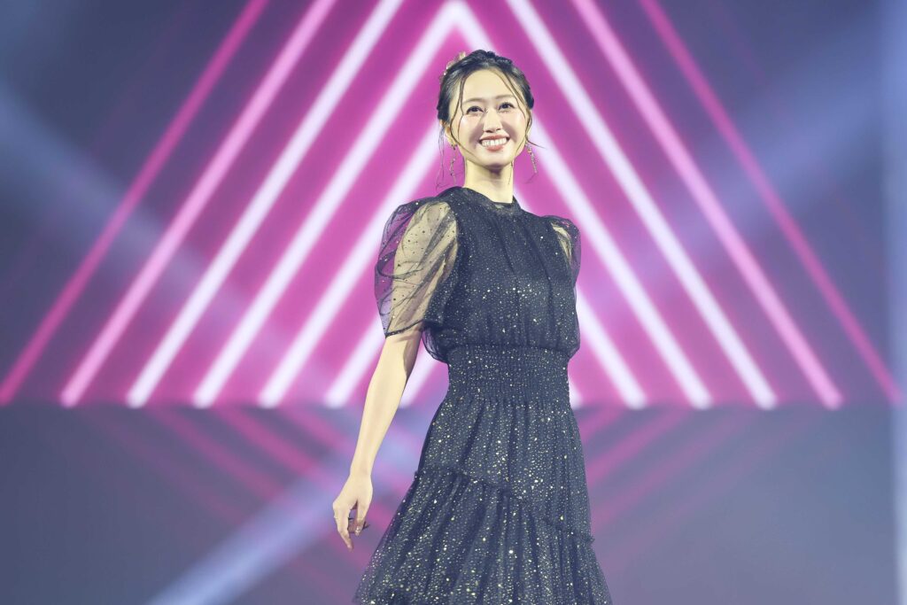 Smiling female performer on stage in a glittery black dress with pink neon lights behind her behind.