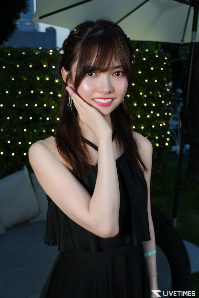 Smiling young woman in a black dress poses outdoors, her hand under her chin with twinkling lights in the background.